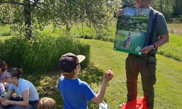 Ein Mann hält ein Poster mit einem Hasenbild hoch, während um ihn herum Kinder auf einer Wiese sitzen. Im Hintergrund ist ein Baum und sonniges Wetter erkennbar.