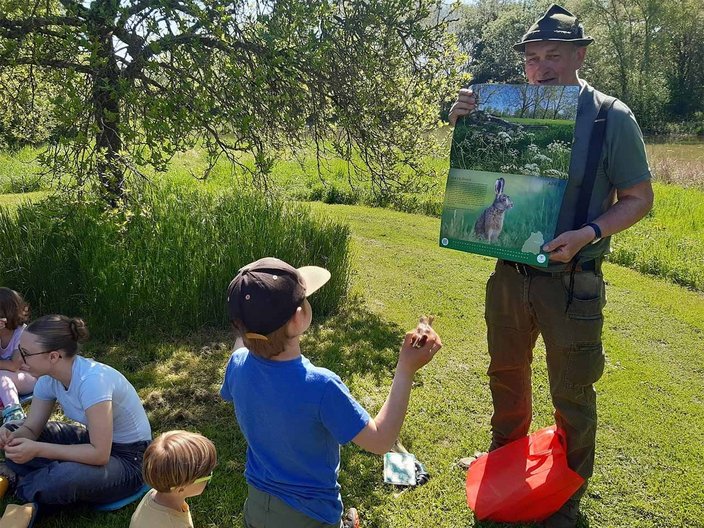 Ein Mann hält ein Poster mit einem Hasenbild hoch, während um ihn herum Kinder auf einer Wiese sitzen. Im Hintergrund ist ein Baum und sonniges Wetter erkennbar.